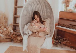 Cozy indoor scene with someone holding a baby in an egg chair beside a piano and dried flowers.
