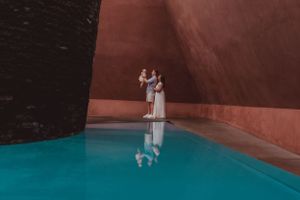 Couple reflected in turquoise pool against terracotta walls.