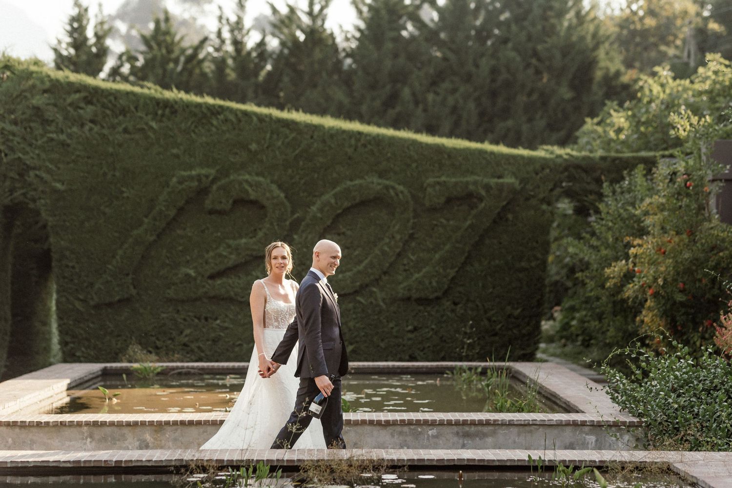Bride and groom lovingly walking hand in hand in front of a serene pond, with a lush green hedge as a tranquil backdrop.