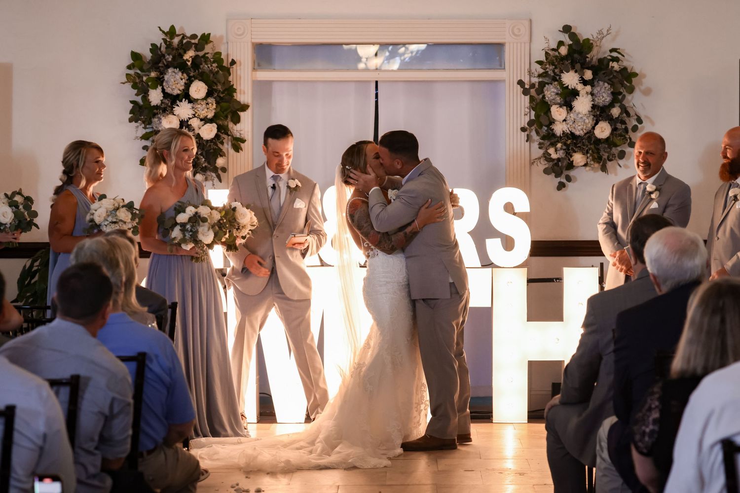 Wedding ceremony moment with illuminated LOVE sign and floral arrangements in background.