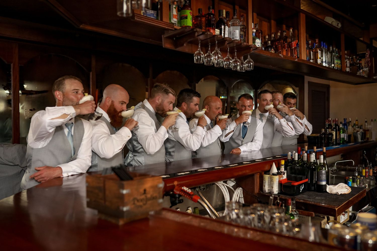 Groomsmen in matching grey vests line up at a bar taking synchronized shots.