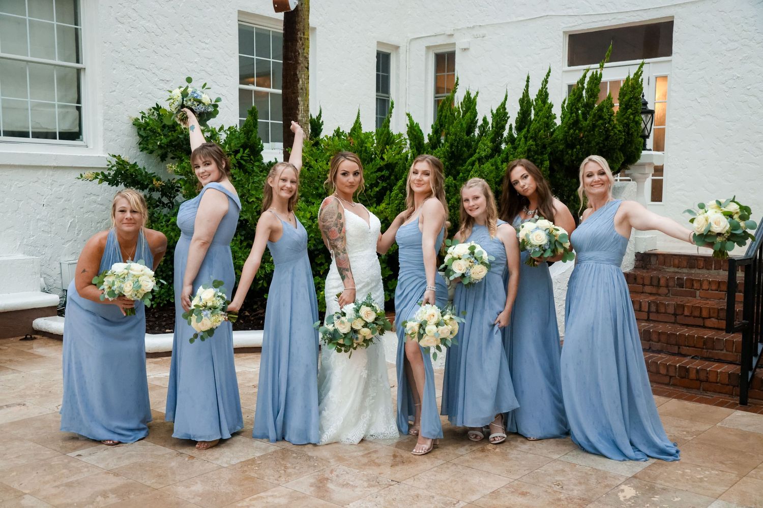 Bridal party in flowing light blue dresses poses with bouquets on outdoor patio.