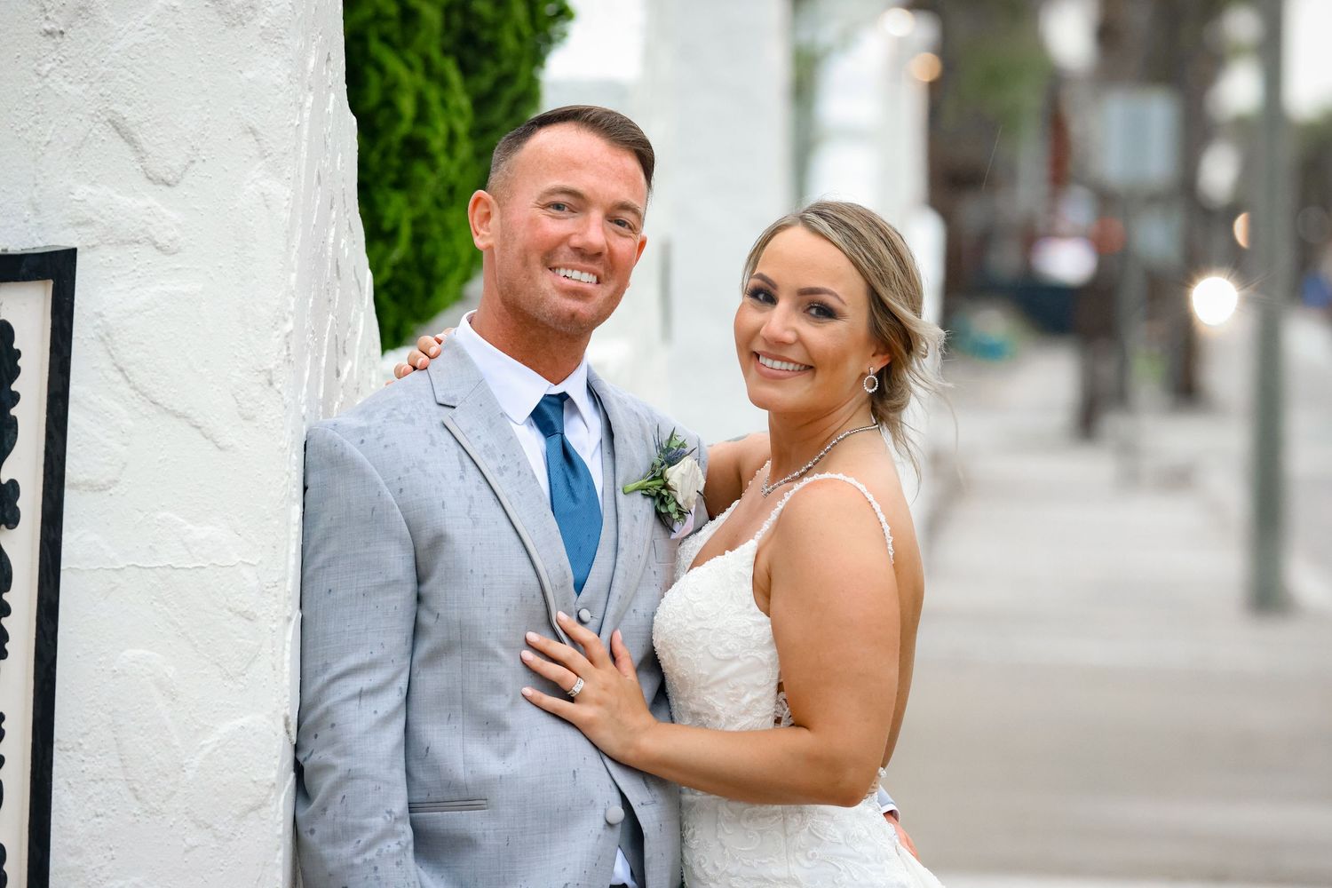 Elegant wedding couple in grey suit and white dress pose against white wall exterior.