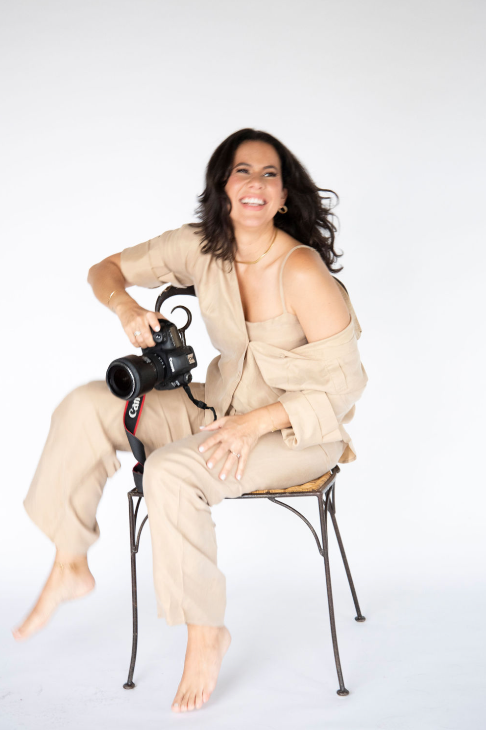 Casual studio portrait with photographer holding camera while seated on metal chair.