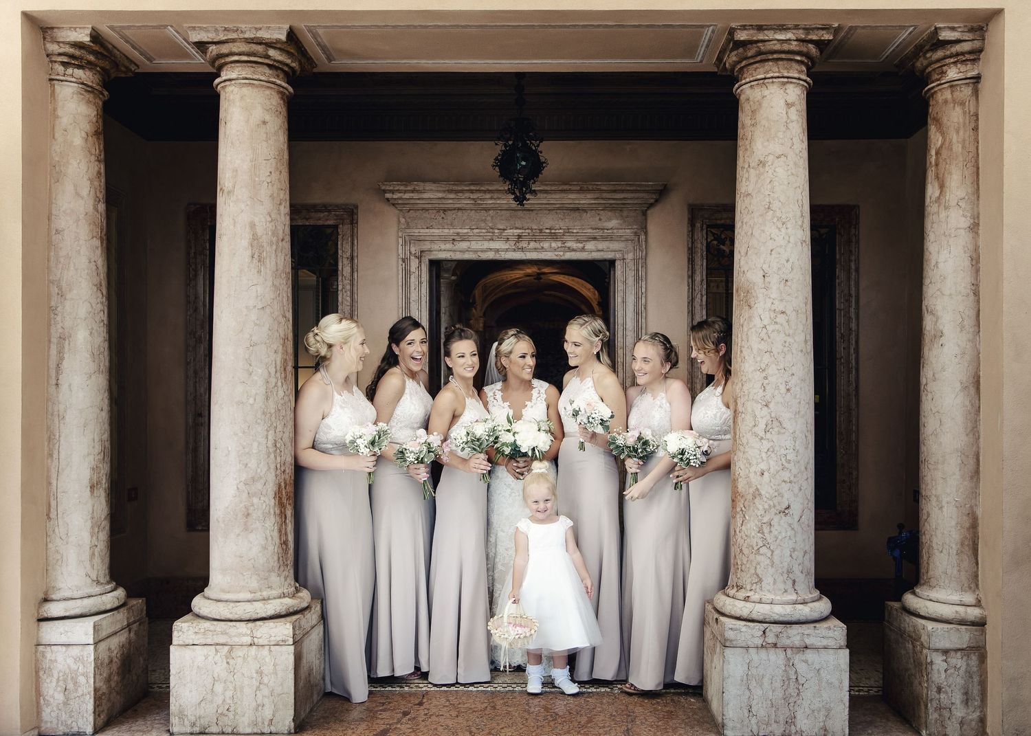 Bridesmaids in light colored dresses pose together in classical colonnade setting.