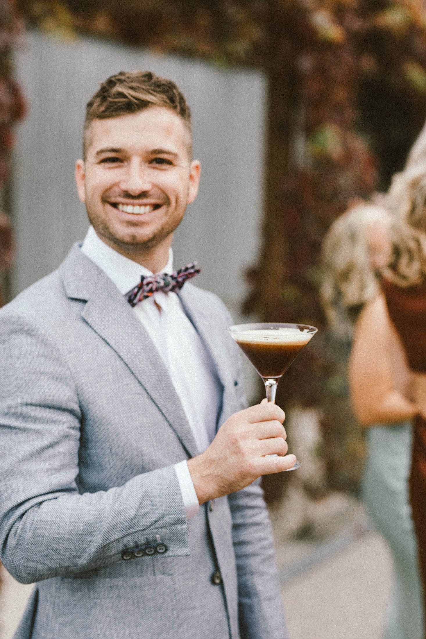 Groomsman in grey suit holding cocktail martini glass during wedding reception.