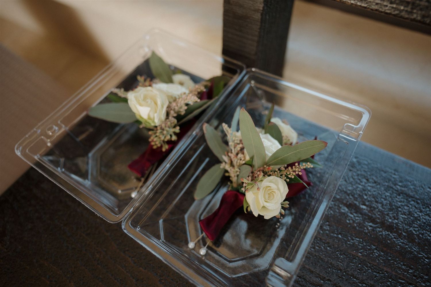Two delicate floral boutonnieres with white roses and greenery in clear plastic boxes.