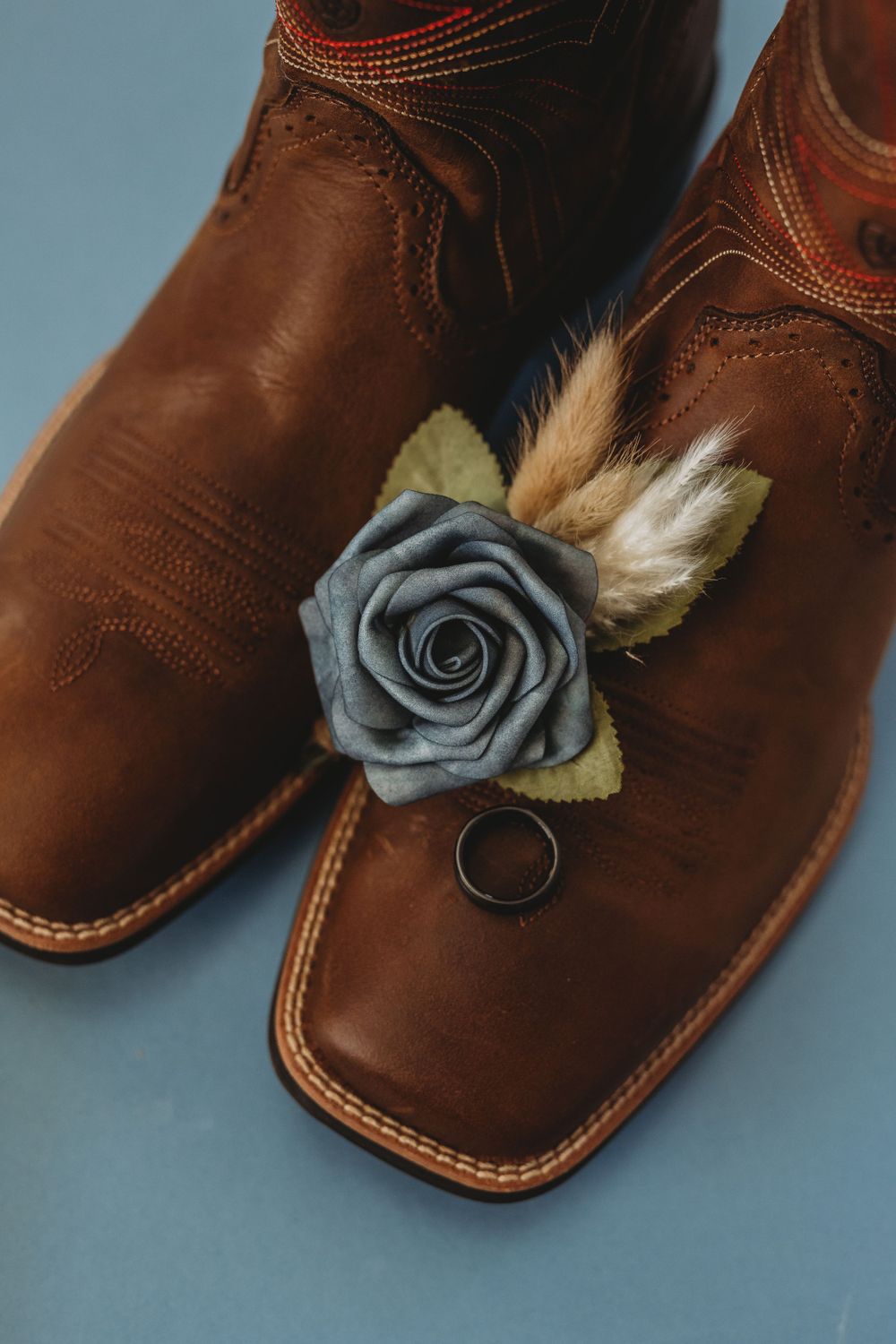 A gray fabric rose boutonniere with dried elements placed on brown leather dress boots.