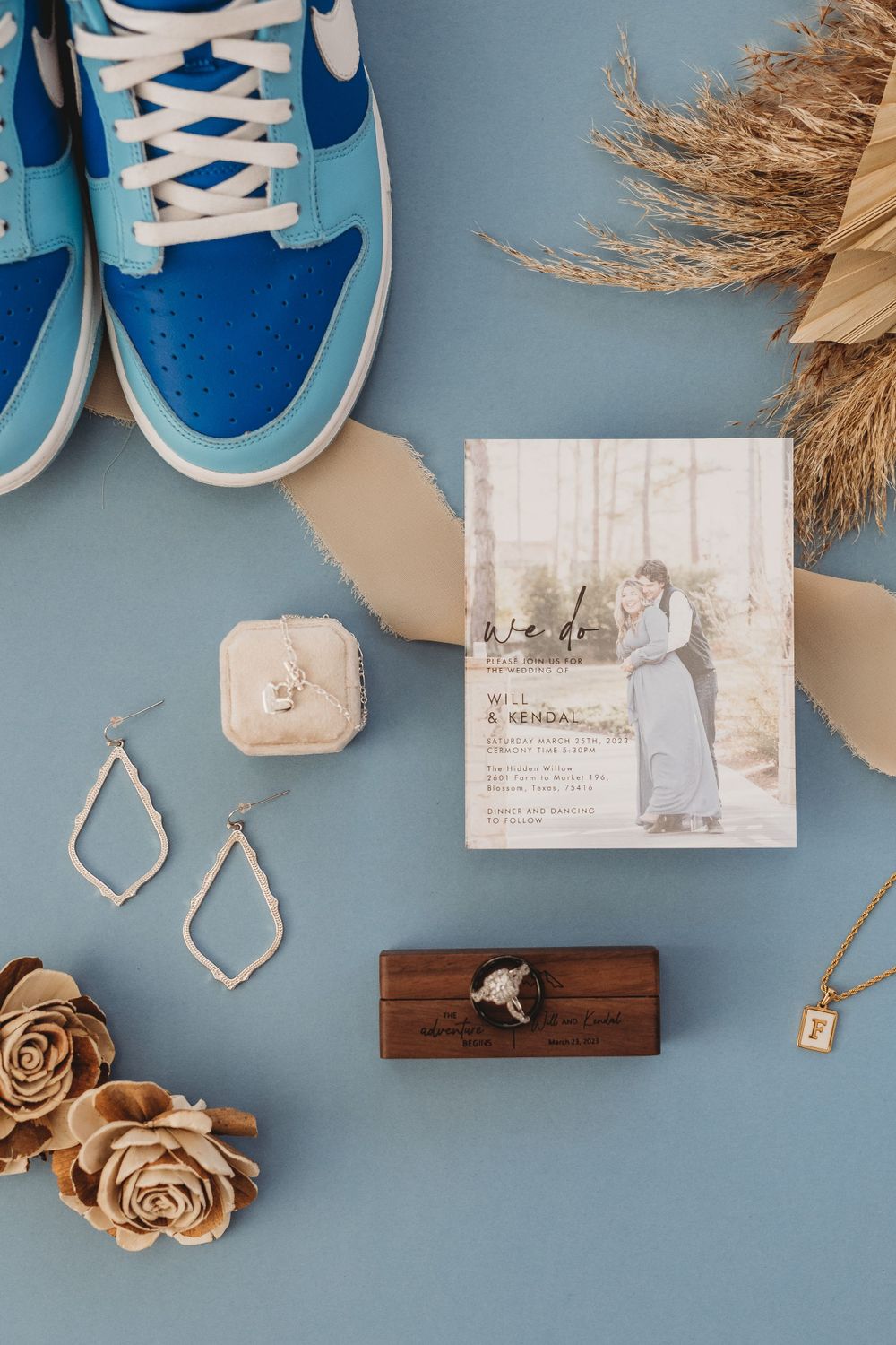 Blue Nike sneakers, jewelry, and wedding invitations styled with dried flowers on blue background.