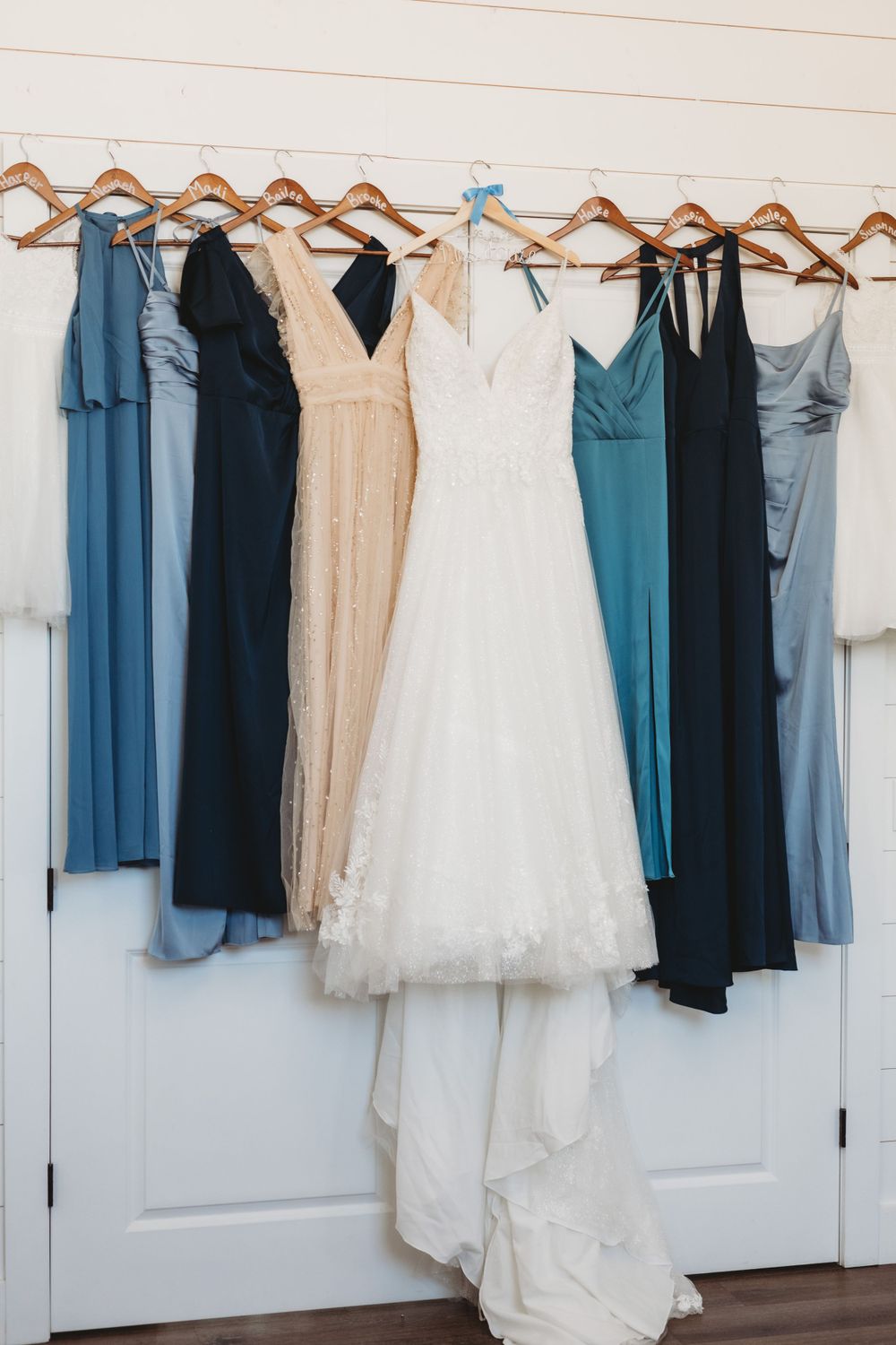Wedding dress and bridesmaid dresses in shades of blue, black, and beige hanging on wooden hangers against a white wall.