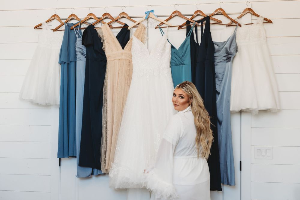 A collection of bridesmaid dresses and wedding gown displayed on wooden hangers against a white wall.