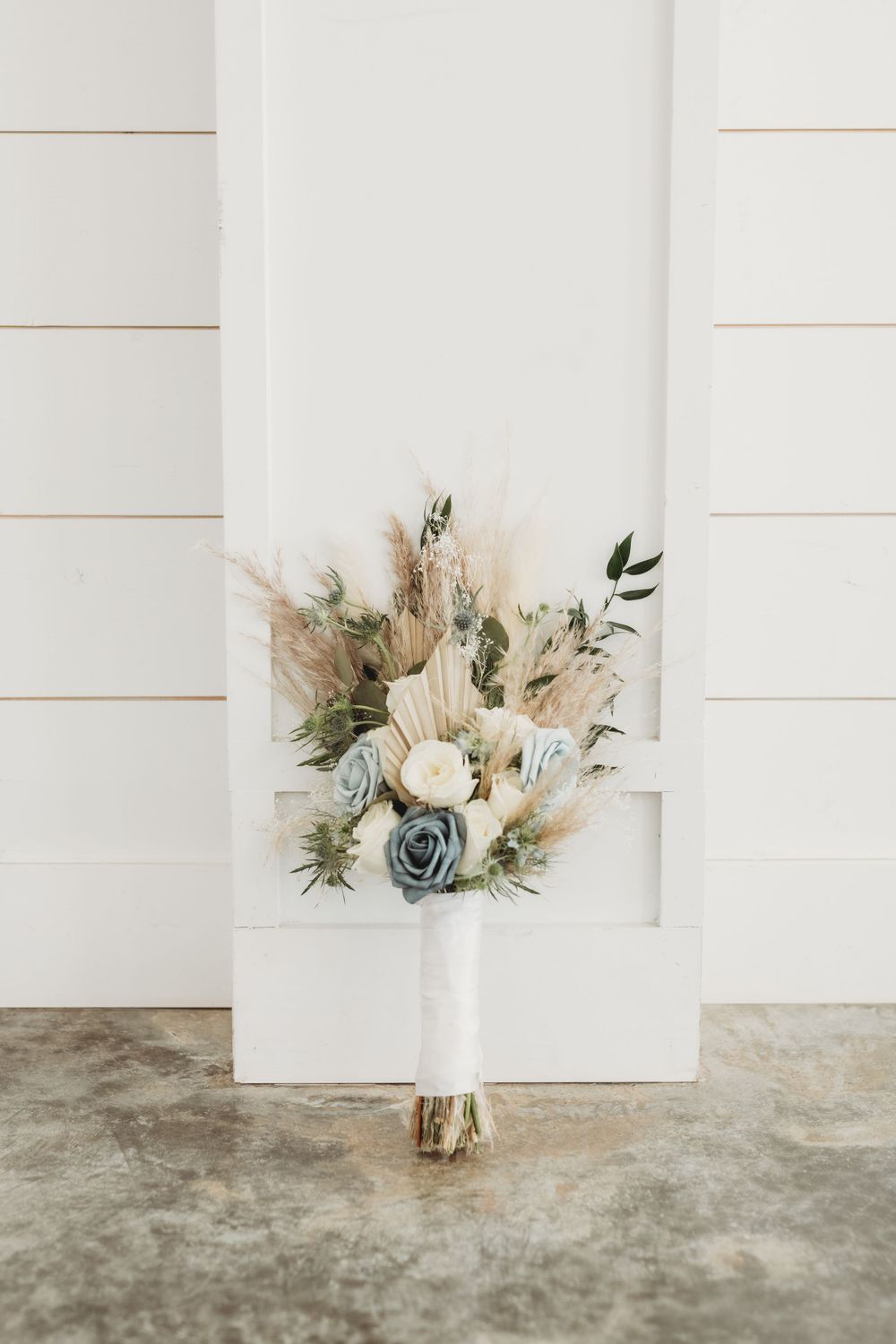 Elegant bridal bouquet with white roses, blue flowers, and pampas grass against a white backdrop.