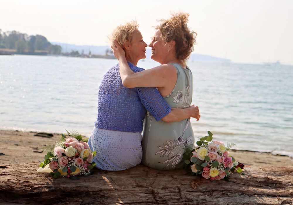 A couple shares an intimate moment on a beach log with floral arrangements.