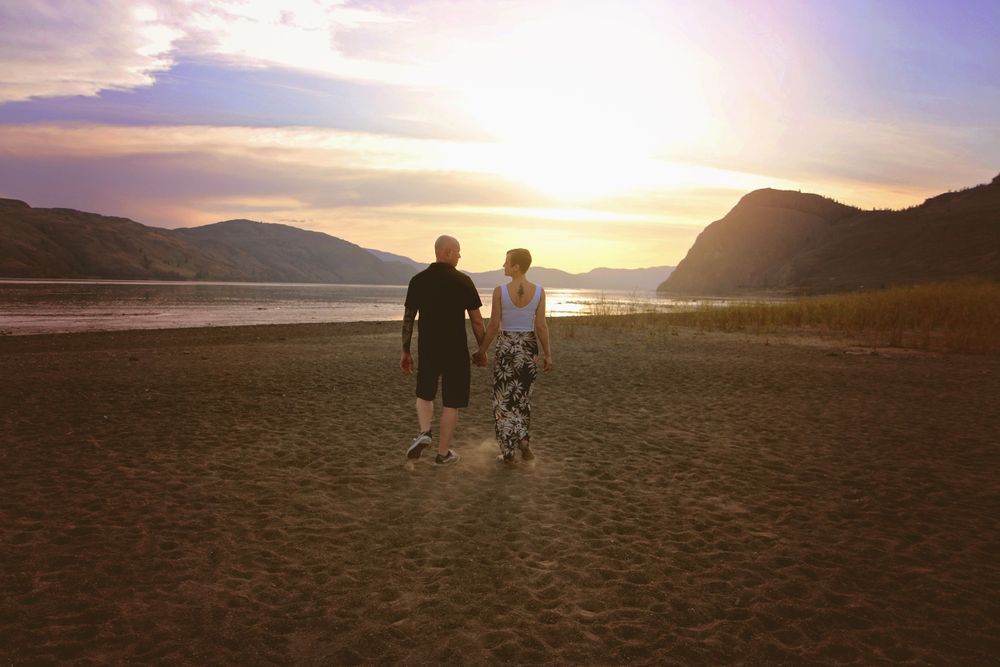 A couple walks hand in hand on a beach at sunset with mountains and water in the background.