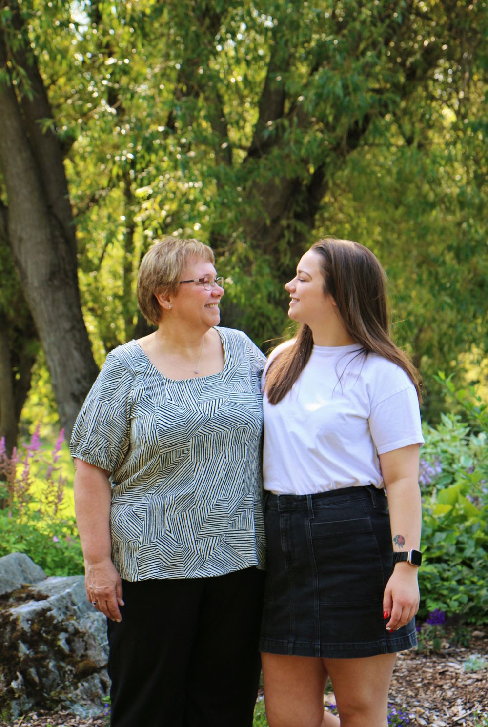 Two people stand close together in a garden setting with lush greenery and summer flowers.