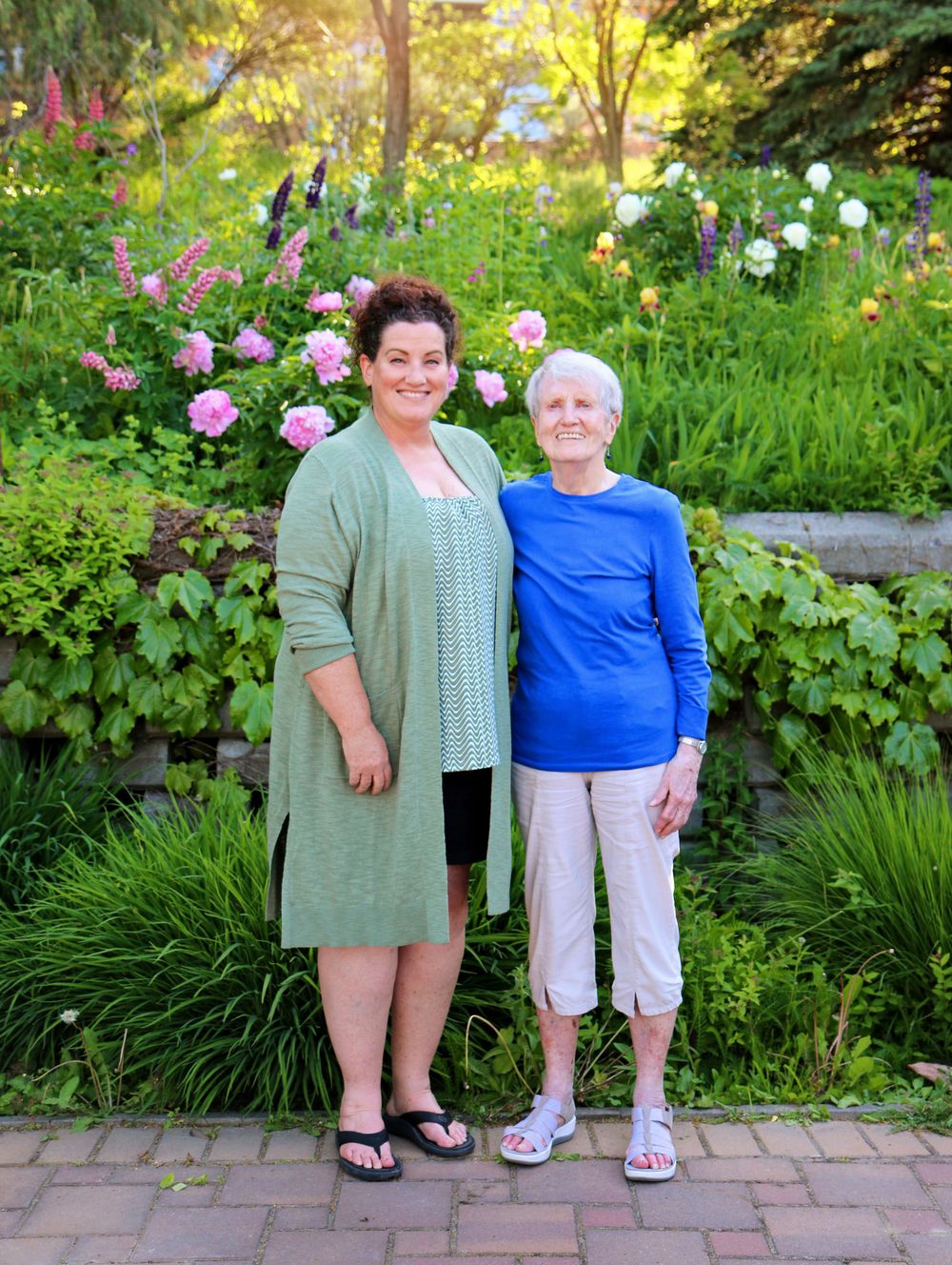 Two people stand together on brick pathway with flowering garden and stone wall behind.