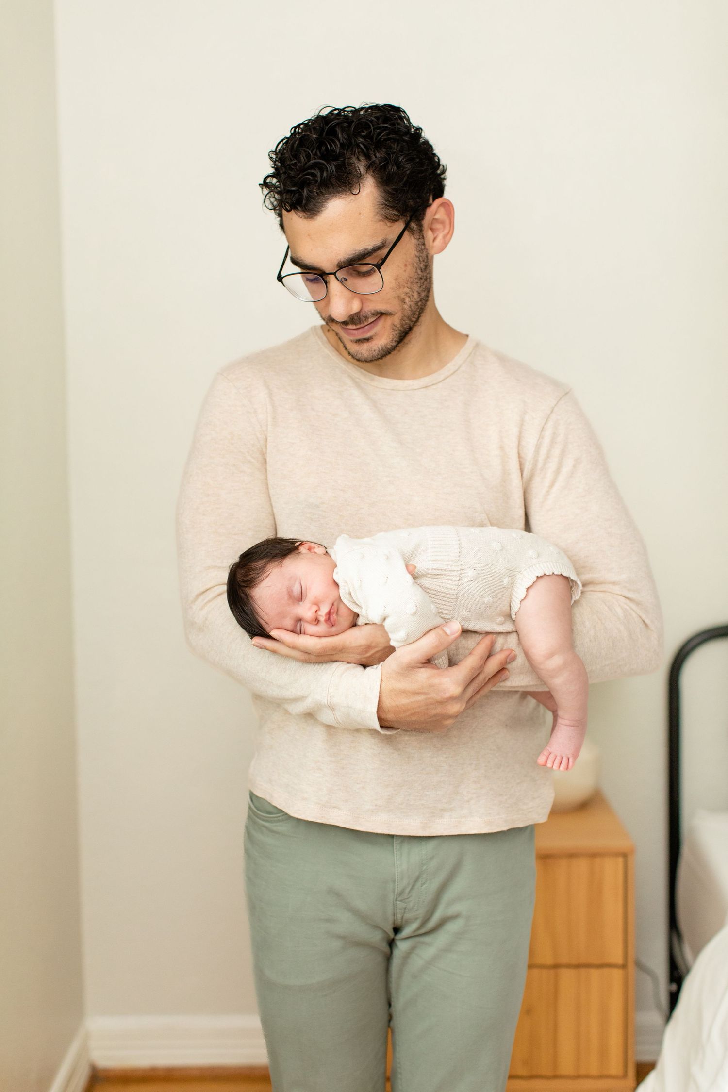 A caregiver in glasses and beige sweater gently holds a sleeping newborn baby.