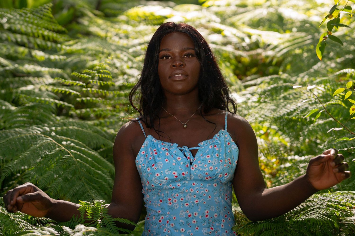 Black woman in blue floral dress poses naturally among lush ferns in sunlit forest.