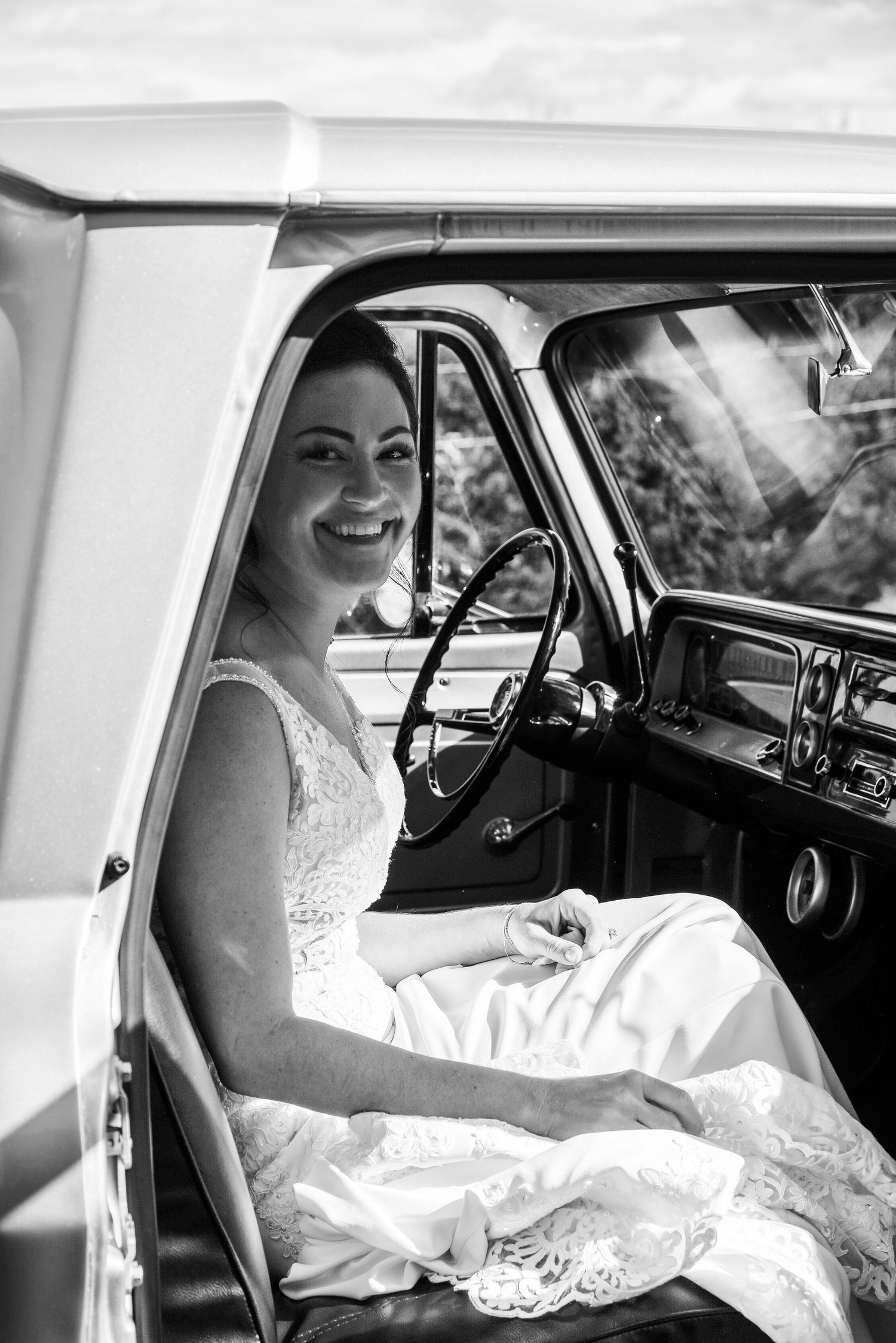 A joyful moment captured in black and white as a bride sits in a vintage car on her wedding day.