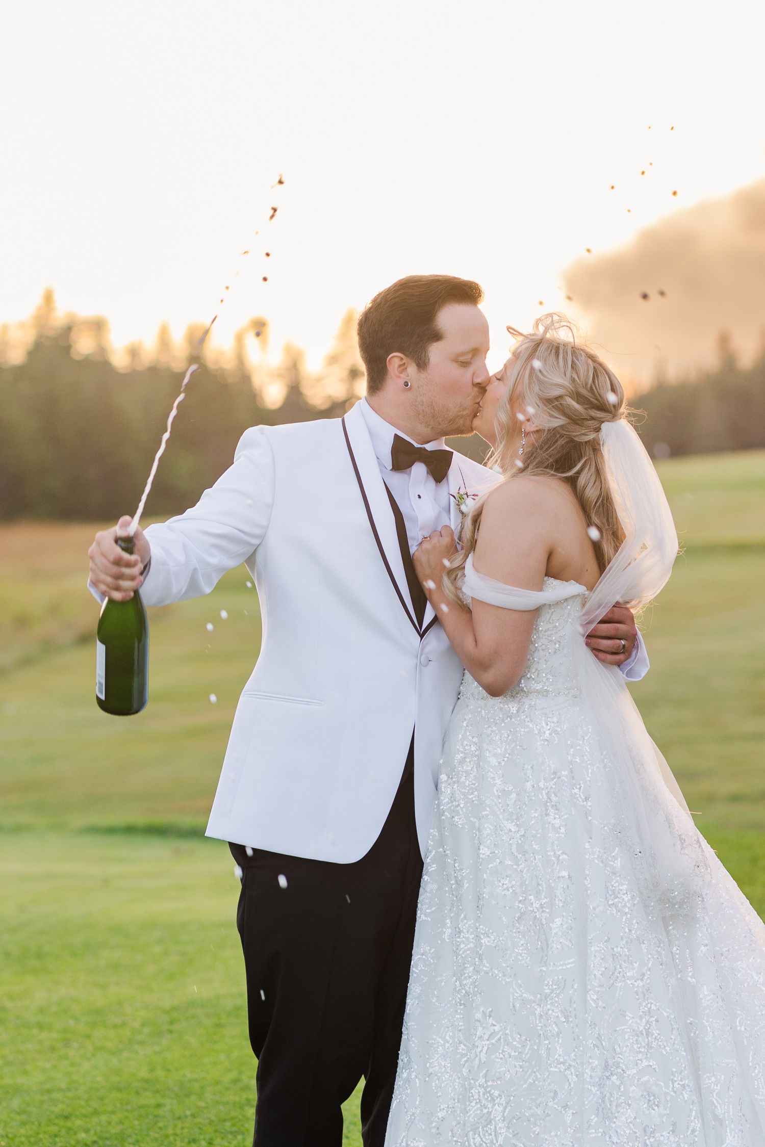 A wedding couple pops champagne and celebrates in a golden field during sunset.