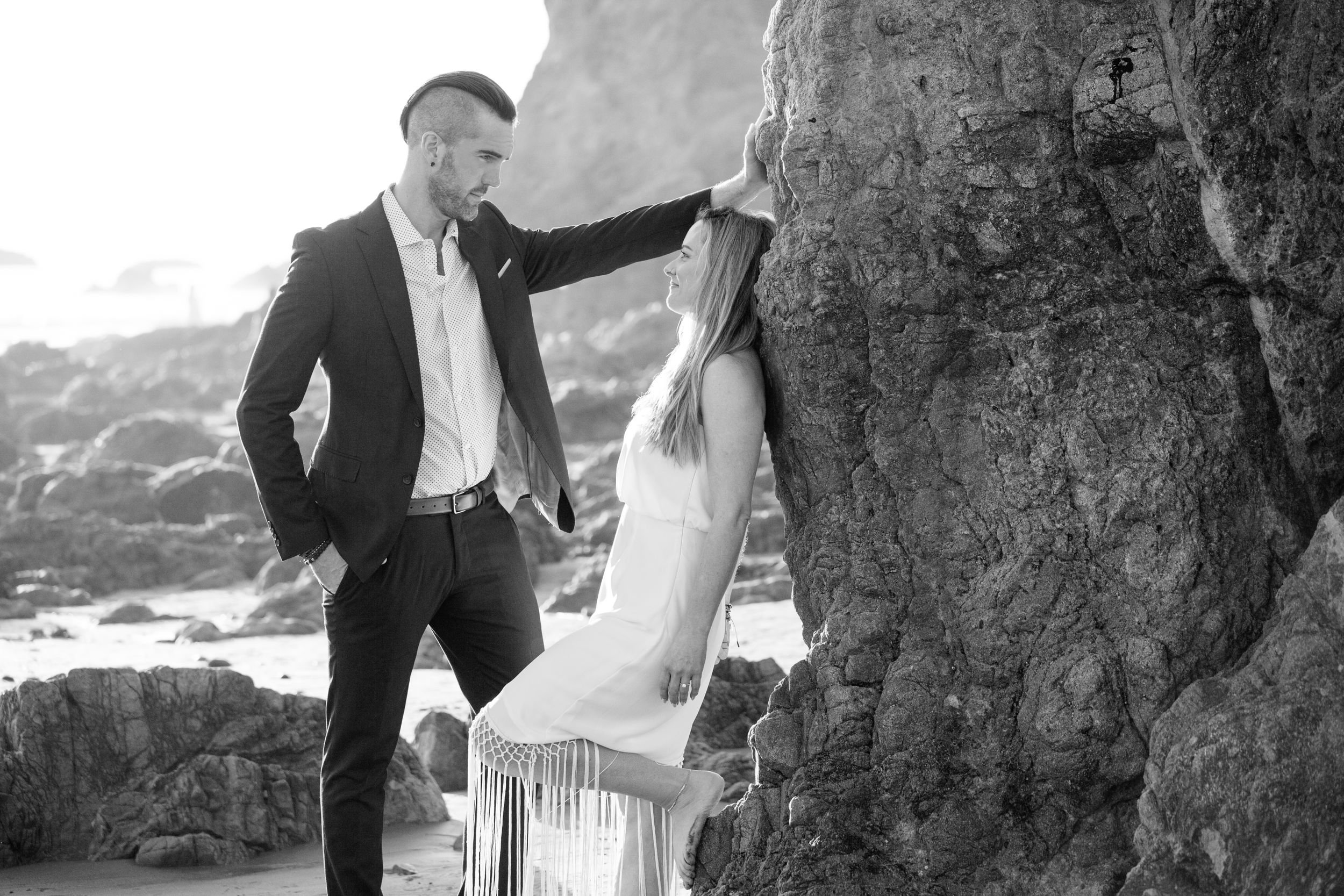 Romantic black and white beach photoshoot featuring a couple posing against dramatic coastal rocks.