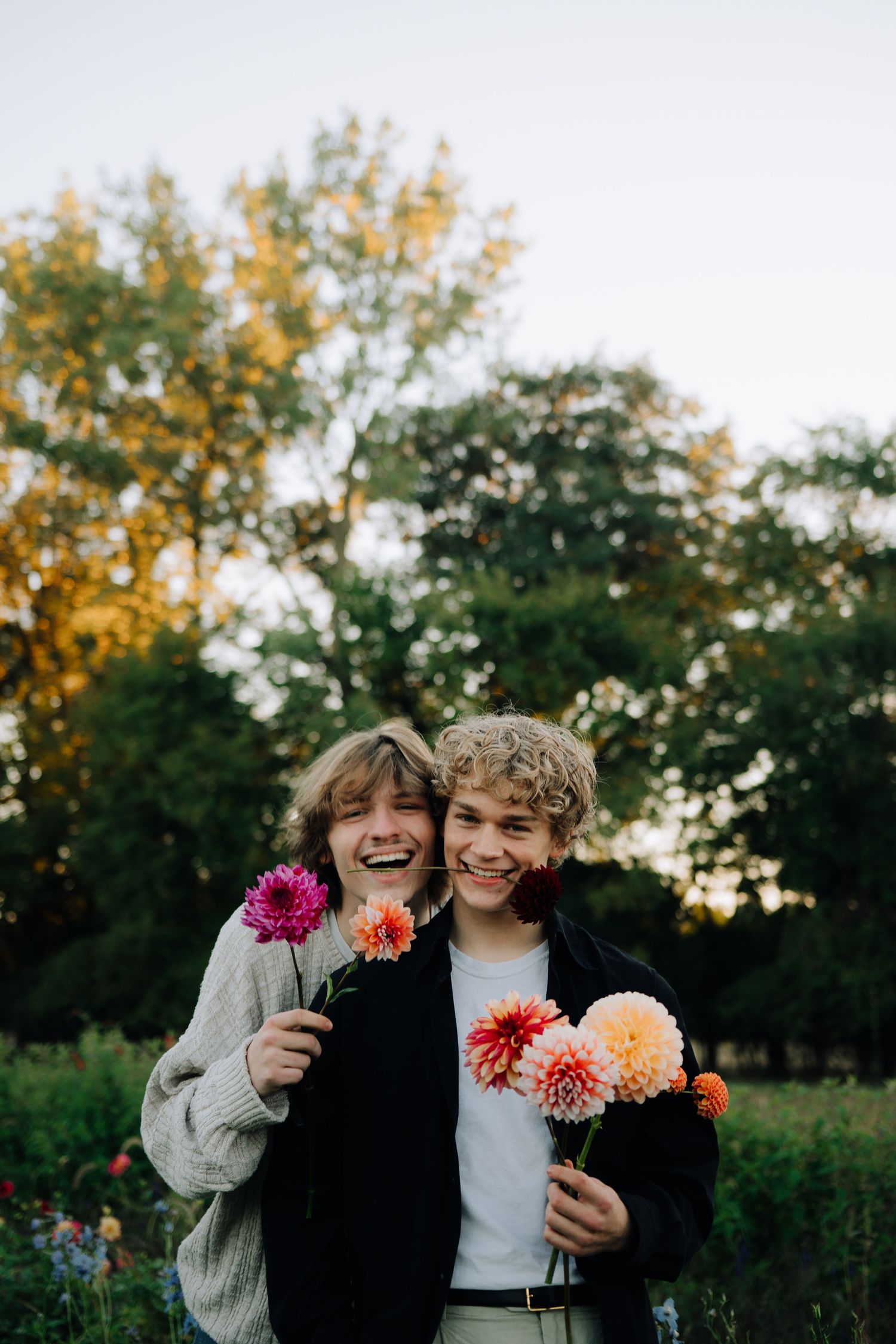 Two friends laughing while holding colorful dahlias in a garden at sunset.