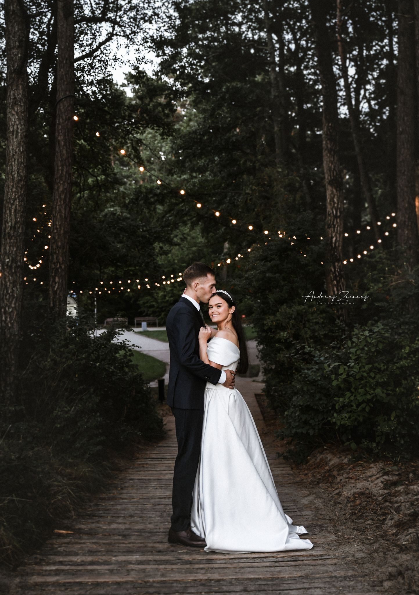 Romantic wedding couple embraces on wooden path under twinkling string lights at dusk.