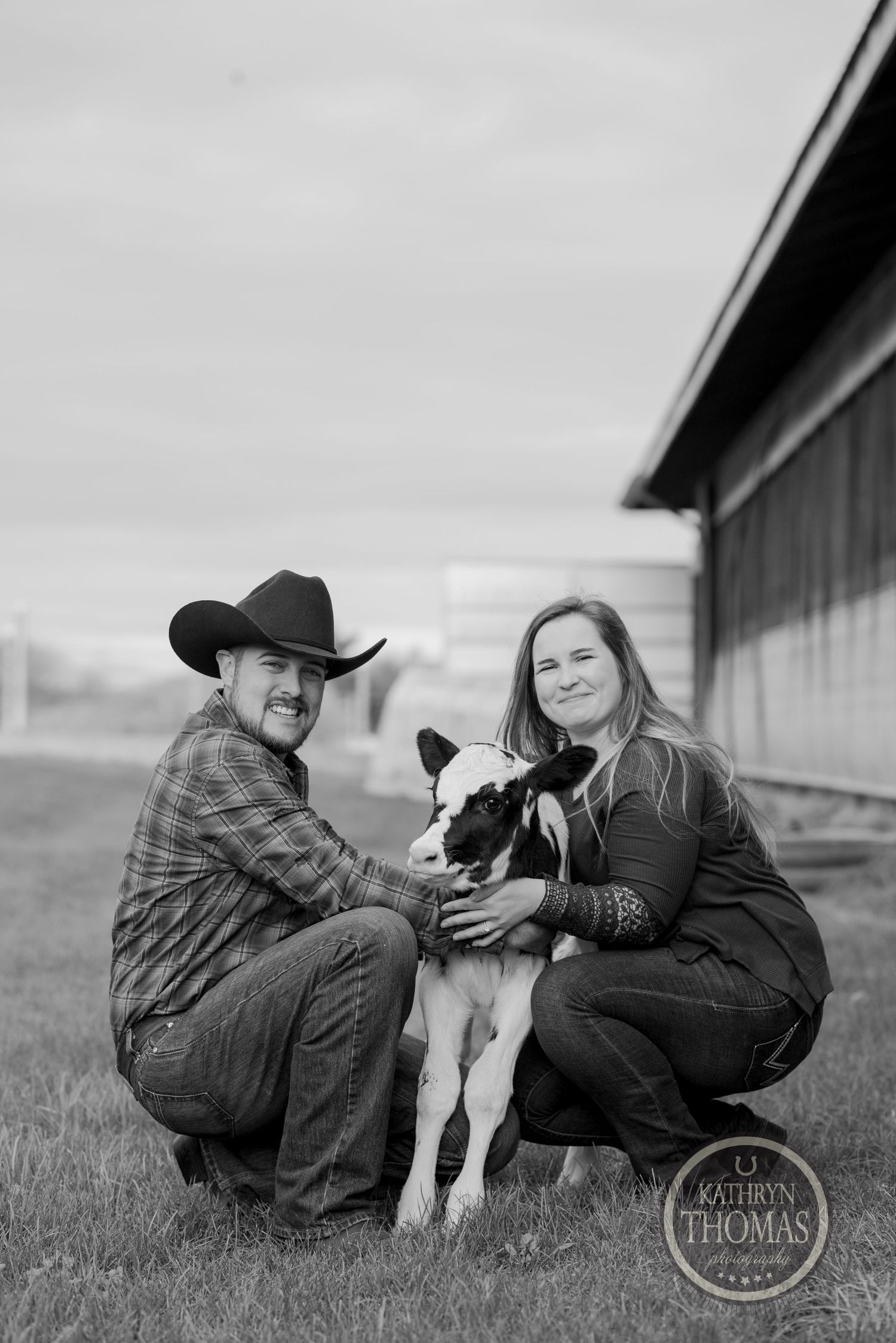 A couple crouches down with their black and white puppy on a farm in a black and white photograph.