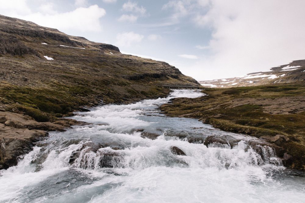 Rushing white water cascades down rocky terrain with patches of snow in a mountainous landscape.