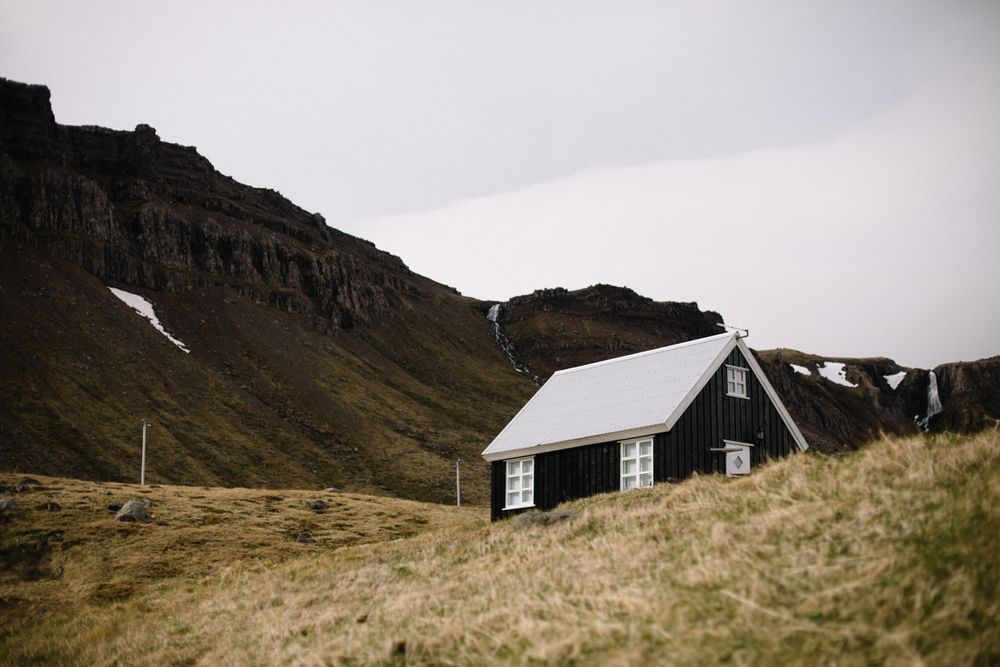 Black wooden cabin nestled against rugged mountain landscape with snow patches.