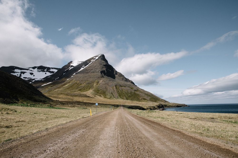 A gravel road leads toward a dramatic mountain peak with snow patches and ocean views in Iceland.