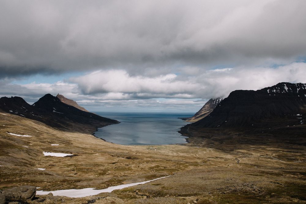 Dramatic fjord landscape with mountains and cloudy skies overlooking a deep blue inlet.