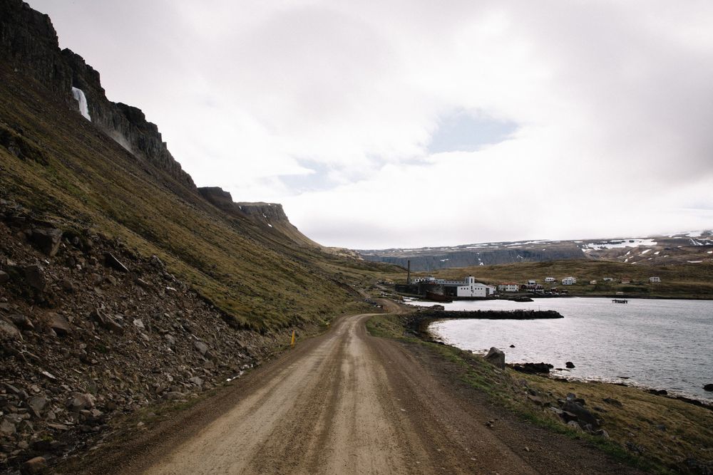 A dirt road winds along a coastal fjord with steep mountain slopes and a boat in the distance.