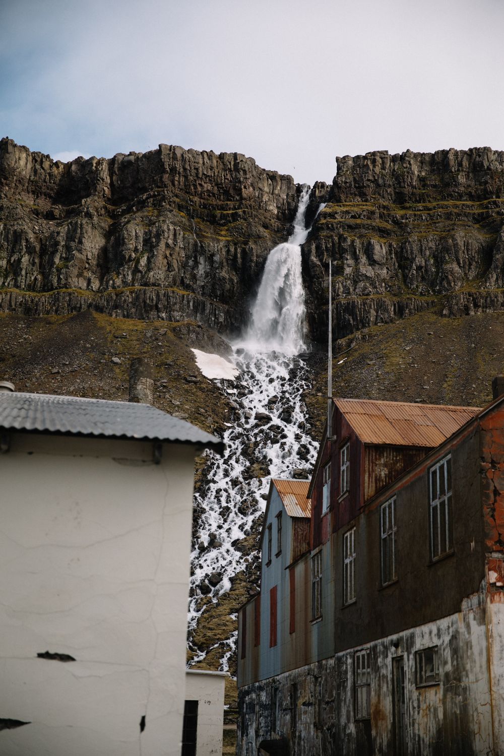 A dramatic waterfall cascades down rocky cliffs behind rustic buildings in a small town.