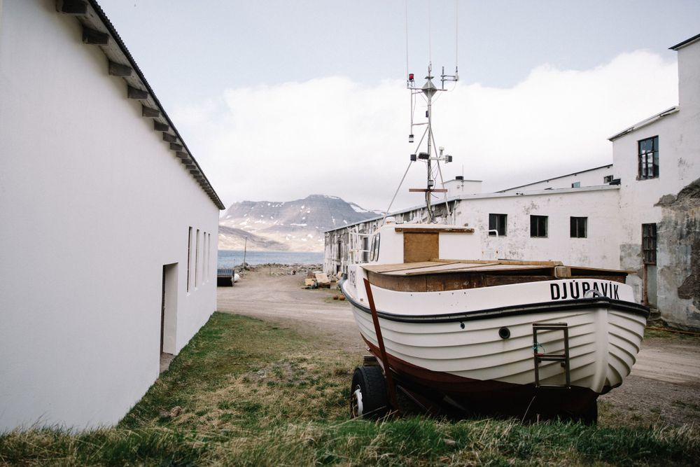 An old fishing boat rests on grass near white buildings with mountains and a communications tower in the background.