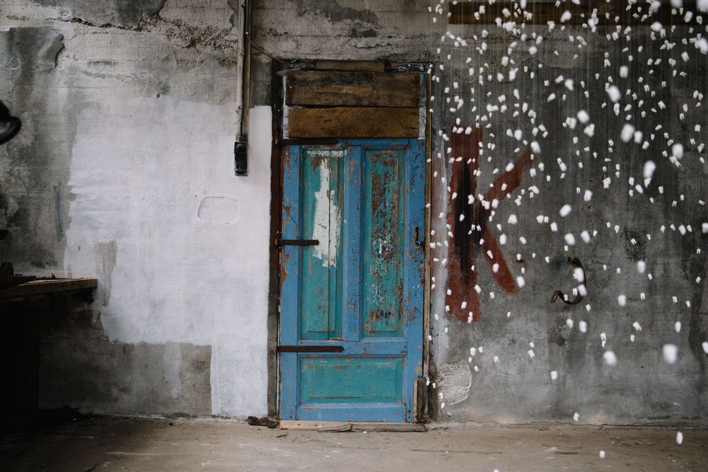 A weathered turquoise blue door stands against a white wall with sparkling snow or light effects in the foreground.