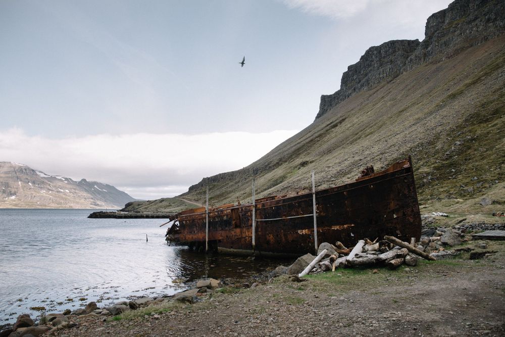 Rustic abandoned boat shelter on rocky coastline with mountains in background.