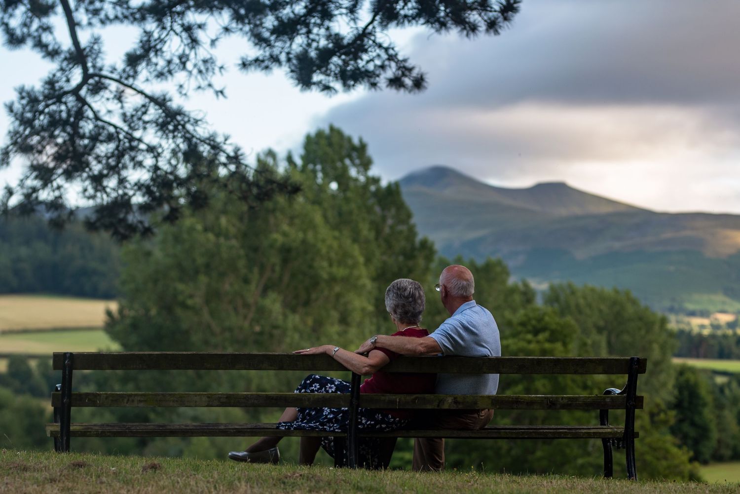 Two people relaxing on a wooden bench with rolling hills and mountains in the background.