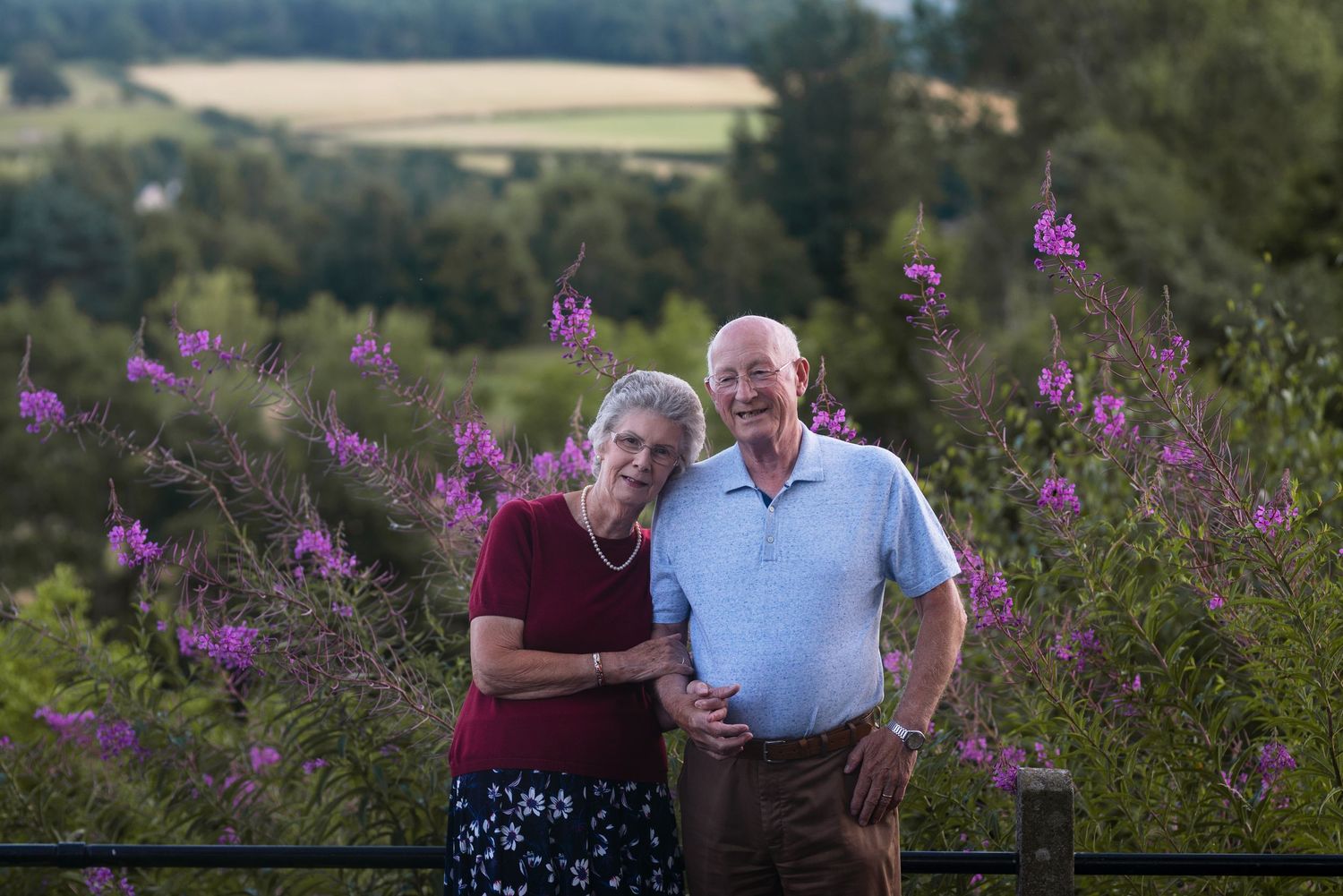 Two people walking together along a countryside path surrounded by blooming purple flowers.