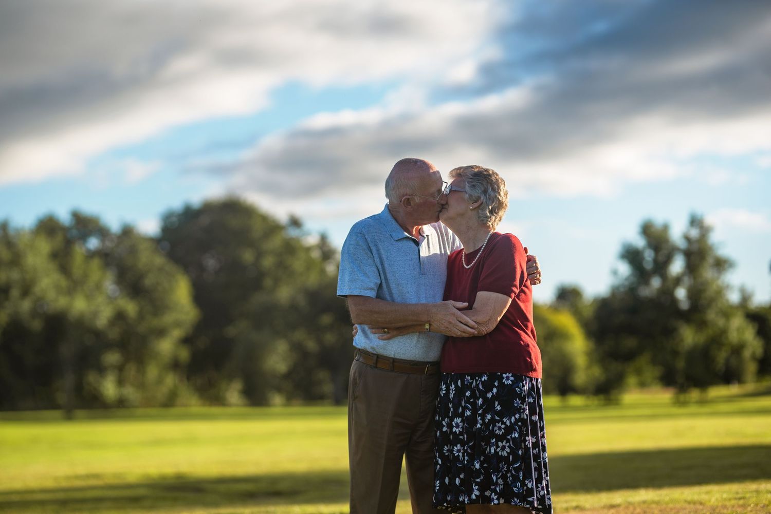 Couple embracing in an open field with blue sky and clouds overhead.