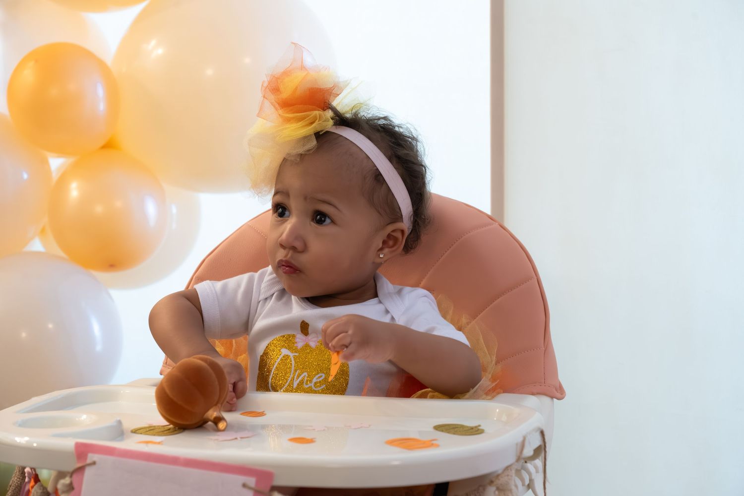 A baby sitting in a high chair with orange balloons in the background during a celebration.