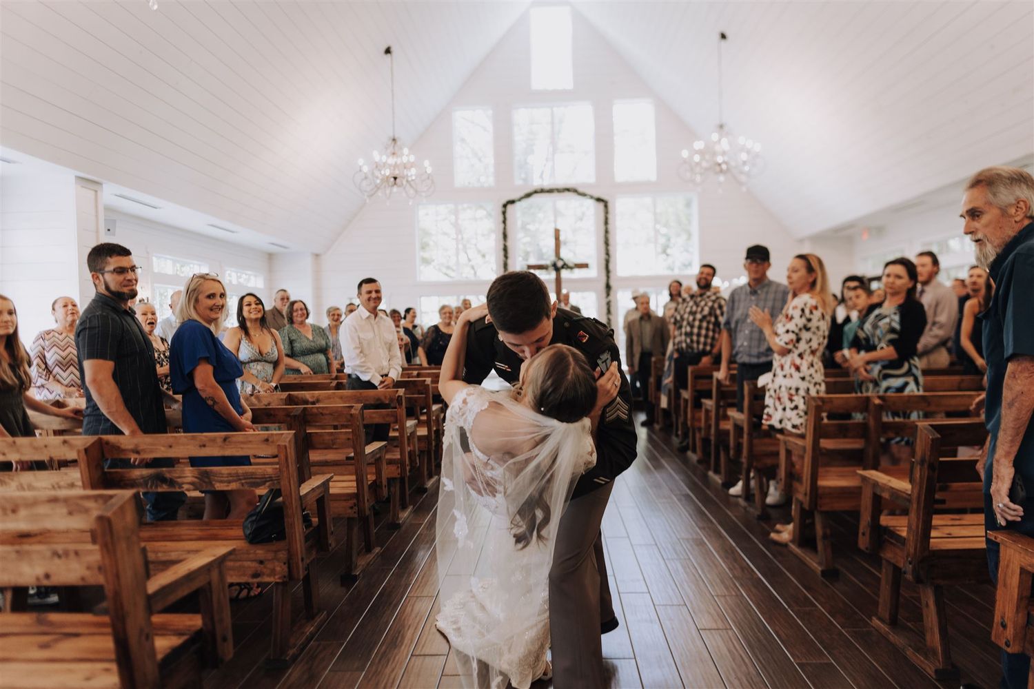 Wedding ceremony in white chapel with wooden pews and gathered guests.