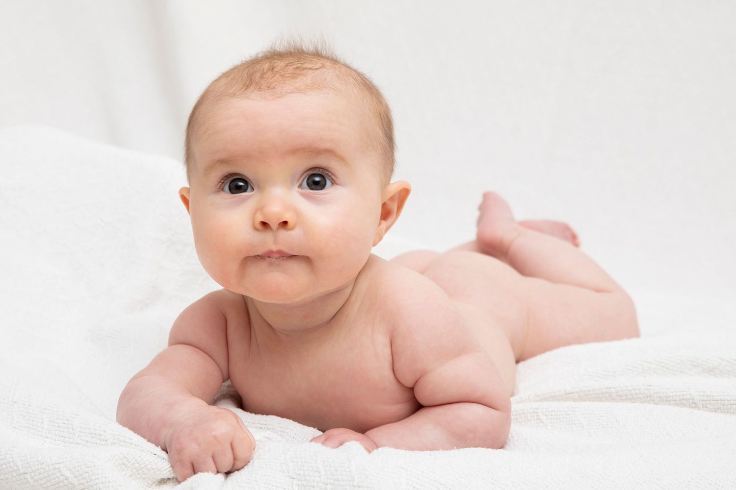 Baby lying on tummy on white blanket looking directly at camera.