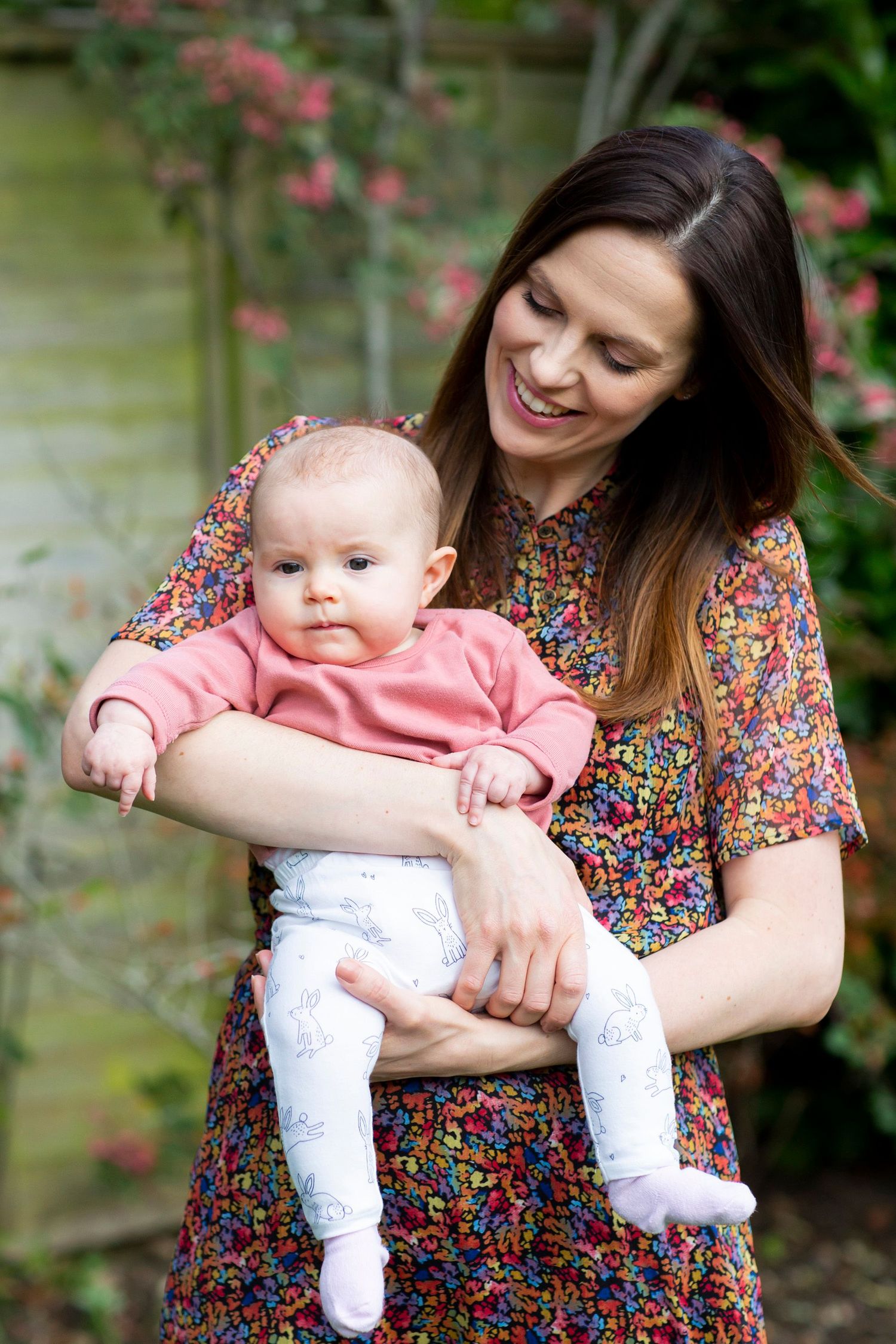 Mother and baby share tender moment in garden with pink flowers in background.