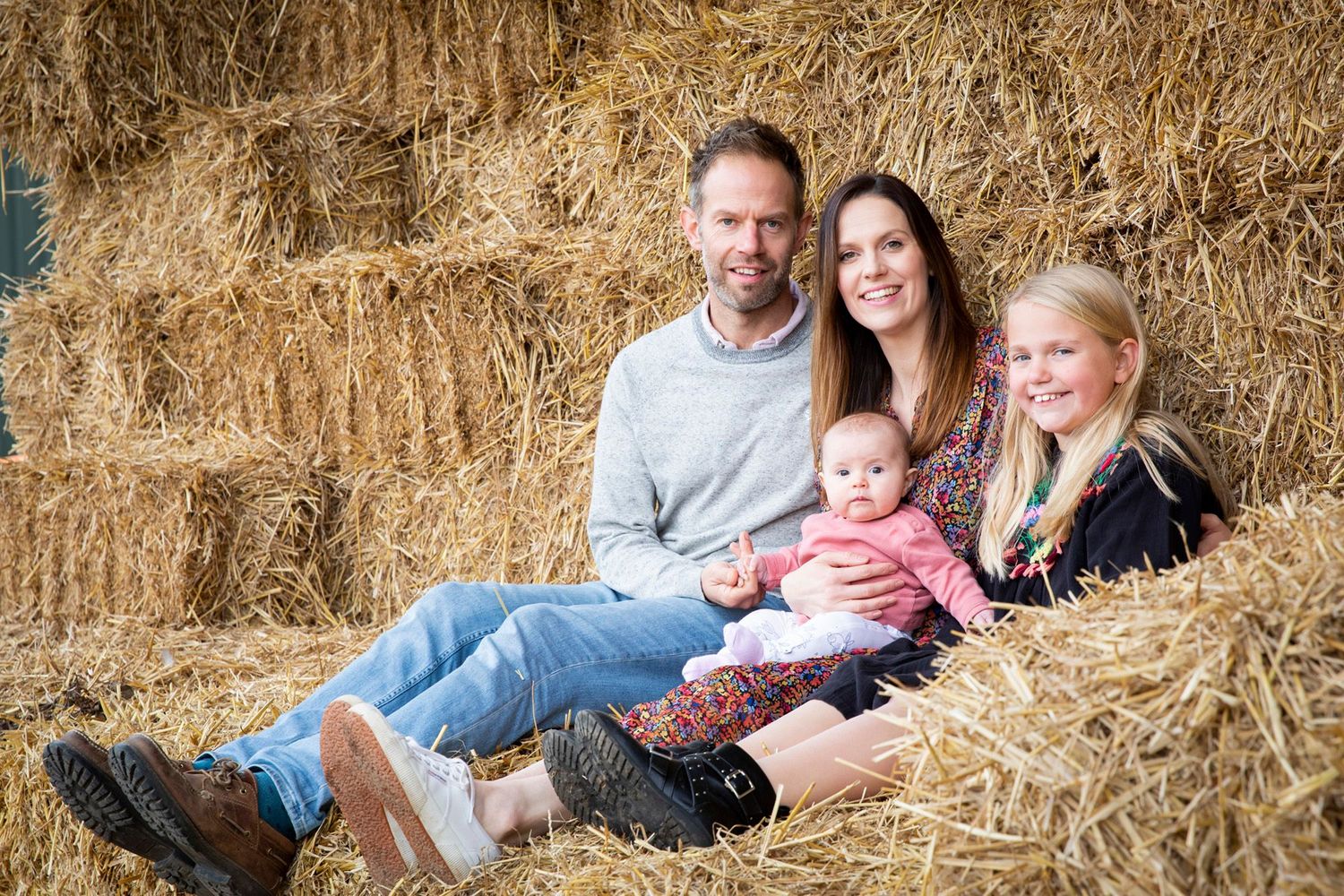 Family sits together in rustic hay bale setting during outdoor photo session.