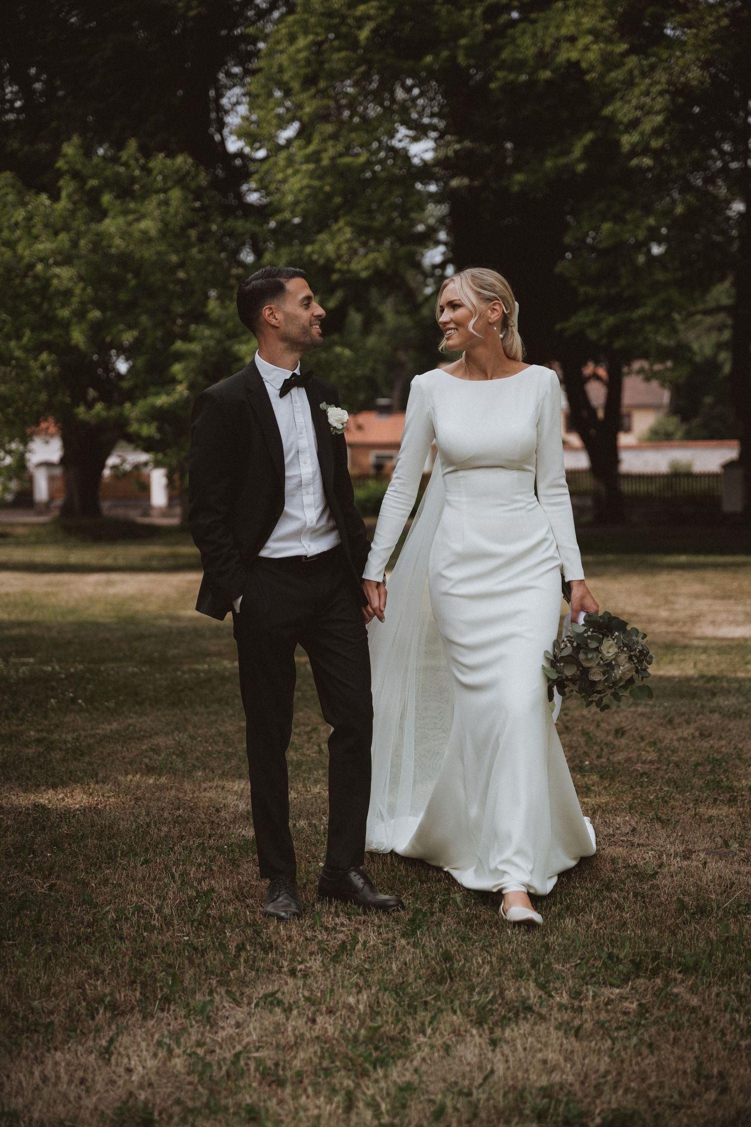 A couple walks together outdoors in formal wedding attire against a natural backdrop.