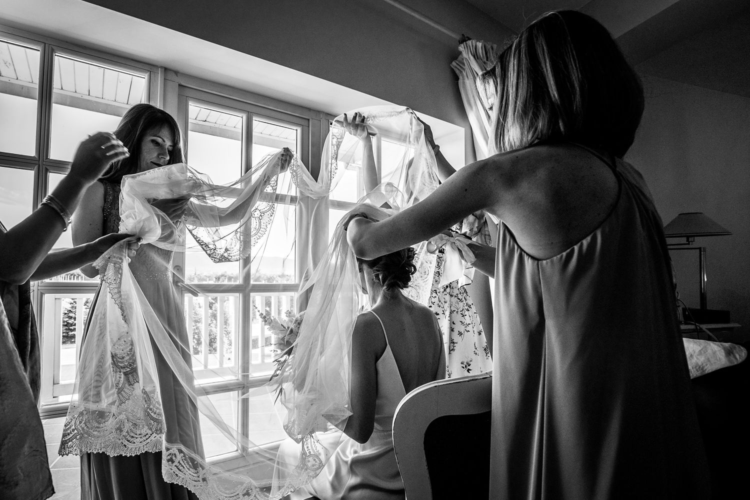 Black and white photo showing preparations on a wedding morning with dress and people getting ready by a window.