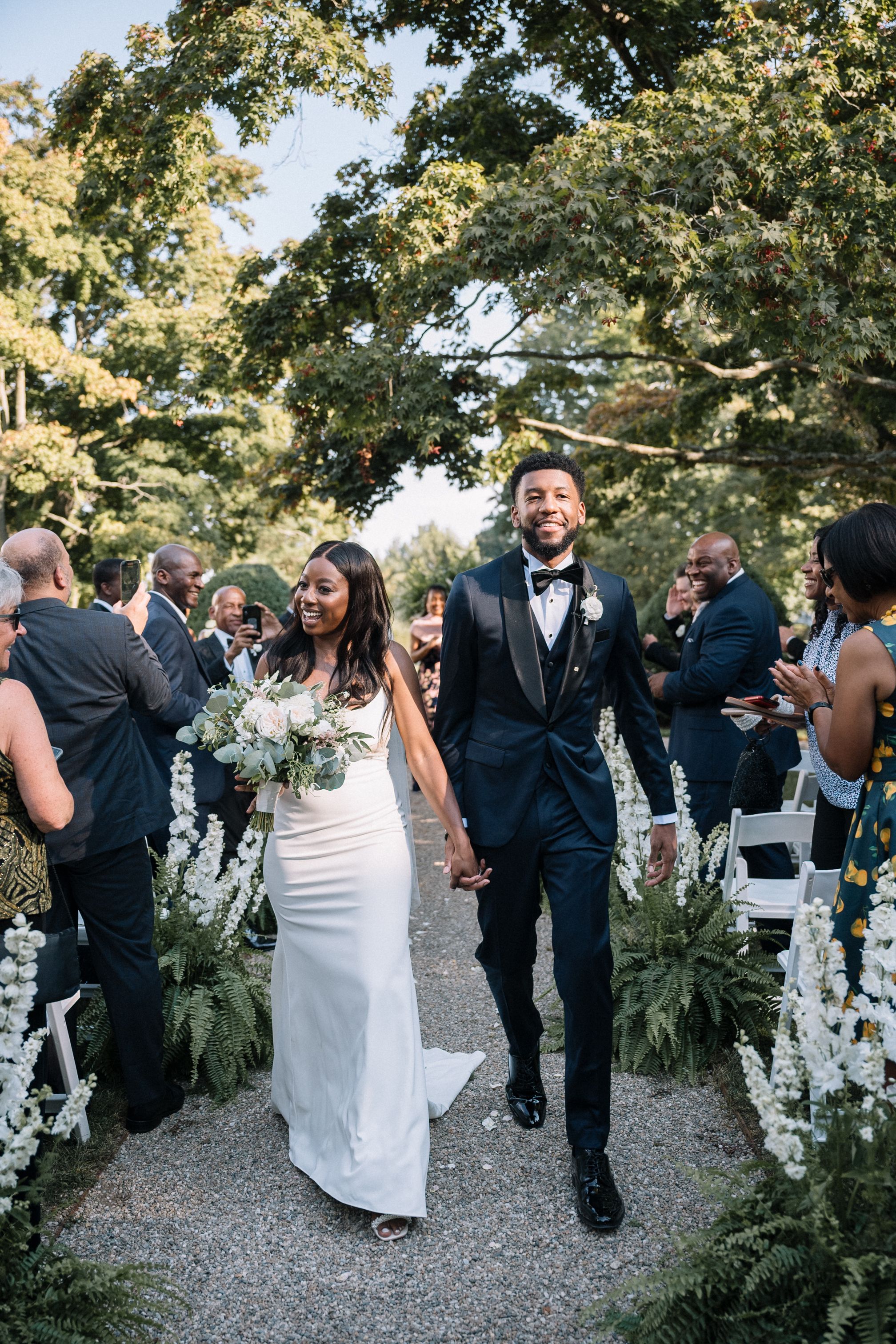 Newlyweds walk down aisle lined with white flowers and greenery as wedding guests celebrate during outdoor ceremony at Burr Mansion