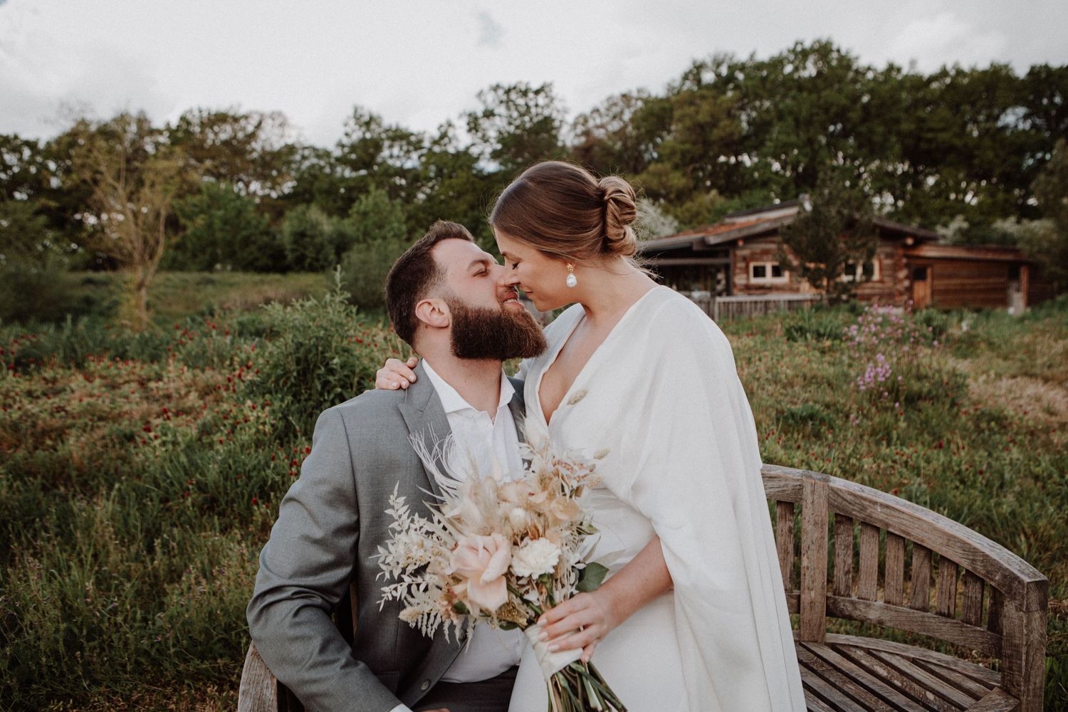 Couple sharing a romantic kiss on a bench surrounded by lush greenery on their wedding day.