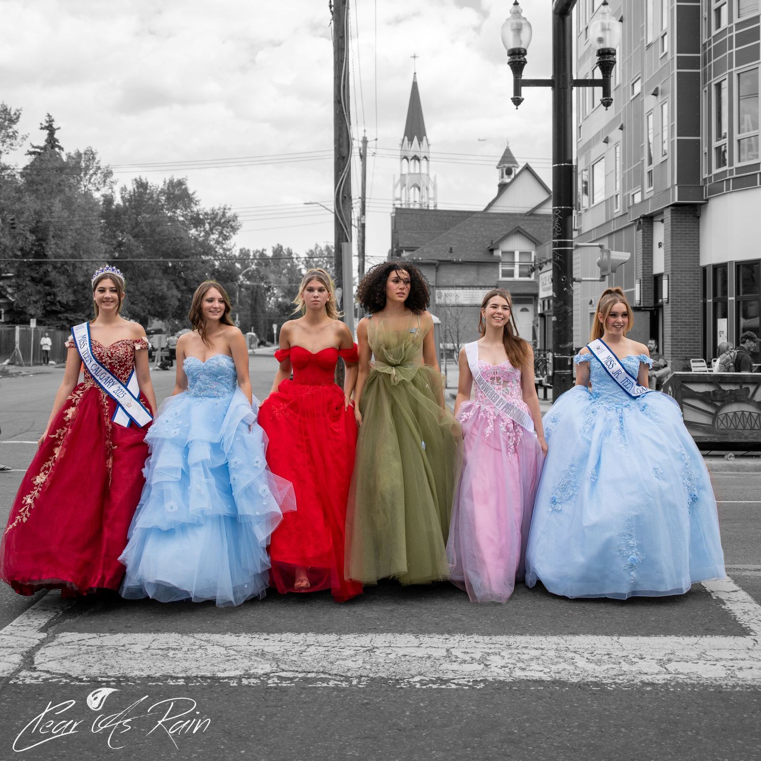 Models in colorful ball gowns pose together on a city street with a church spire visible in the background.